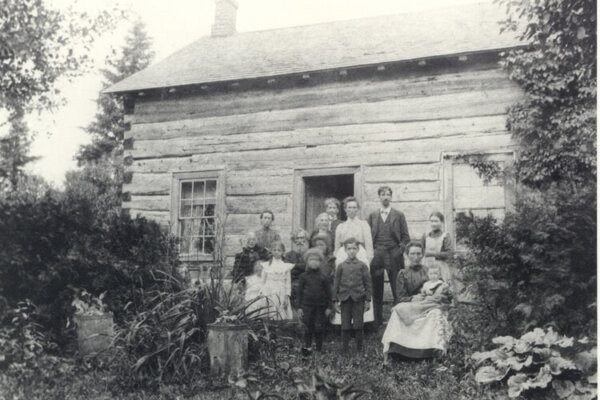 A black and white photograph of a family of 10 people. Standing in front of a rustic log cabin. The log cabin has two windows, a door and chimney.