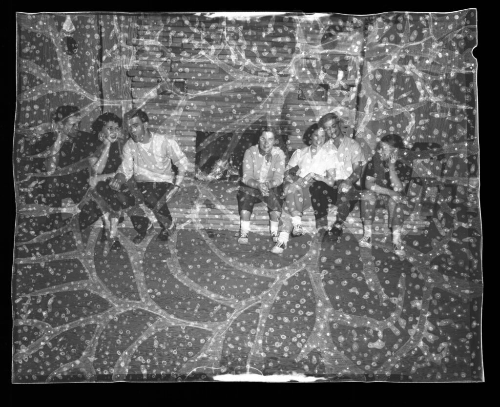 badly deteriorated photograph negative with tiny bubbles across surface. Picture is of couples sitting on the ledge in front of a fireplace.