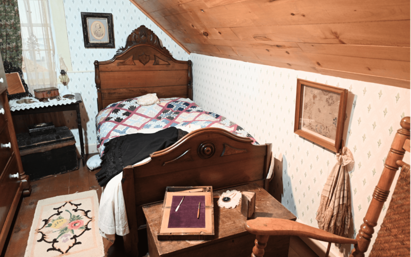 A photograph of a dark wood bed with a diamond patterned quilt on top. Various knick-knacks are spread throughout the room as if someone living there has just placed them down. A cross-stitched piece in a frame is displayed on the wall. A floral rug is on the ground.