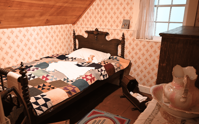 A dark wood bed with a multicoloured quilt on top. In the corner of the room is a pink and white wash basin. On the floor is a wooden doll basinet and a multicoloured rug.