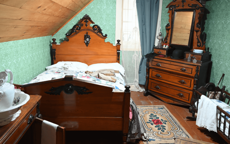 A large bed with a quilt, a wash basin the corner, a crib and mirrored chest of drawers,